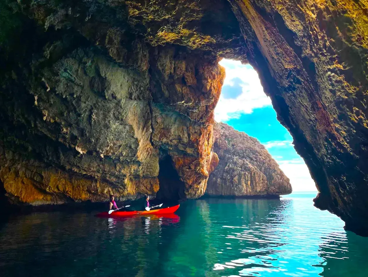 Dos personas en kayak rojo exploran la cueva de la cala del llop mari, con aguas turquesas y formaciones rocosas impresionantes.