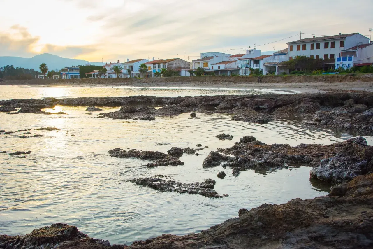 Costa con casas blancas y palmeras, rocas volcánicas en primer plano y el mar reflejando la luz del atardecer. Se divisa una torre la sal a lo lejos.