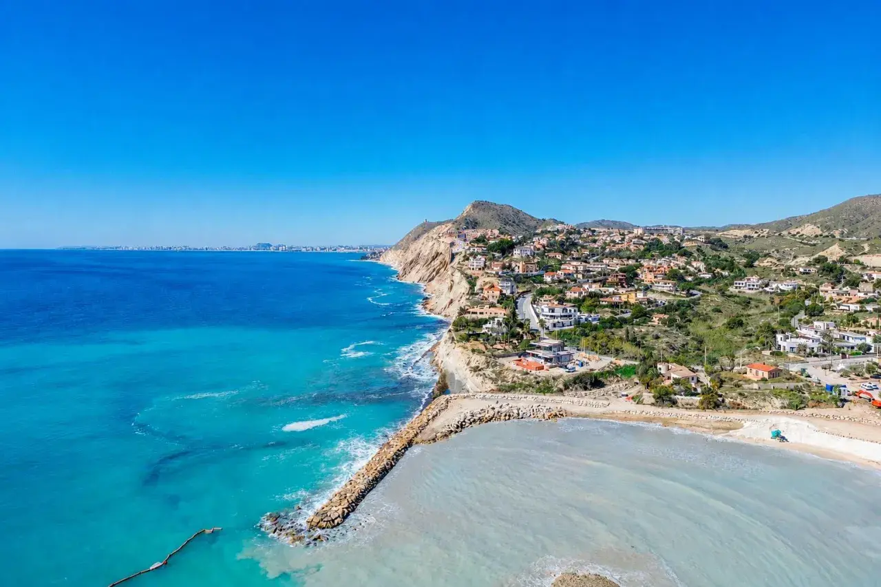Vista aérea de la costa con casas blancas y el mar azul turquesa. La cala Baeza se extiende hacia el horizonte, con una bahía protegida por un rompeolas de rocas.