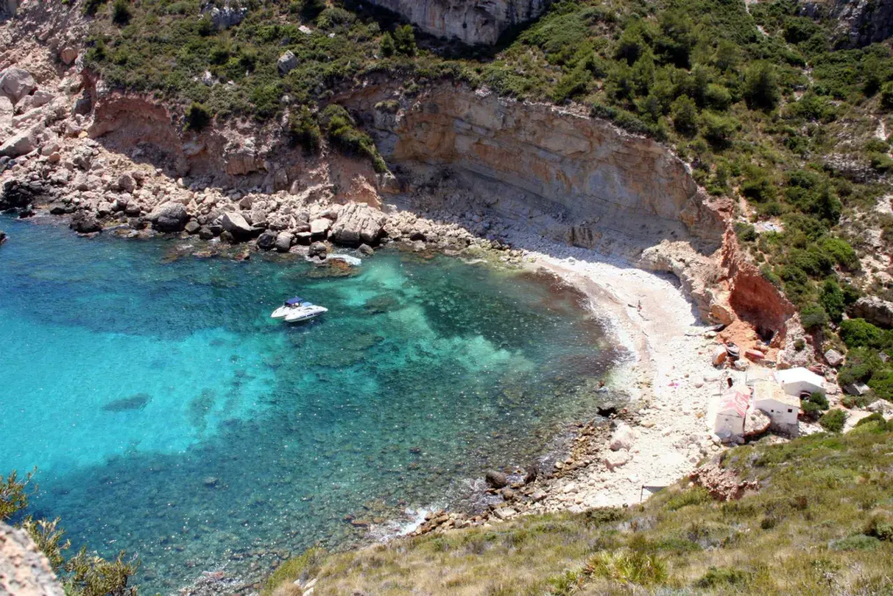 Aguas turquesas y una cala rocosa en Cala Llebeig, con un pequeño barco y casitas blancas en la orilla.