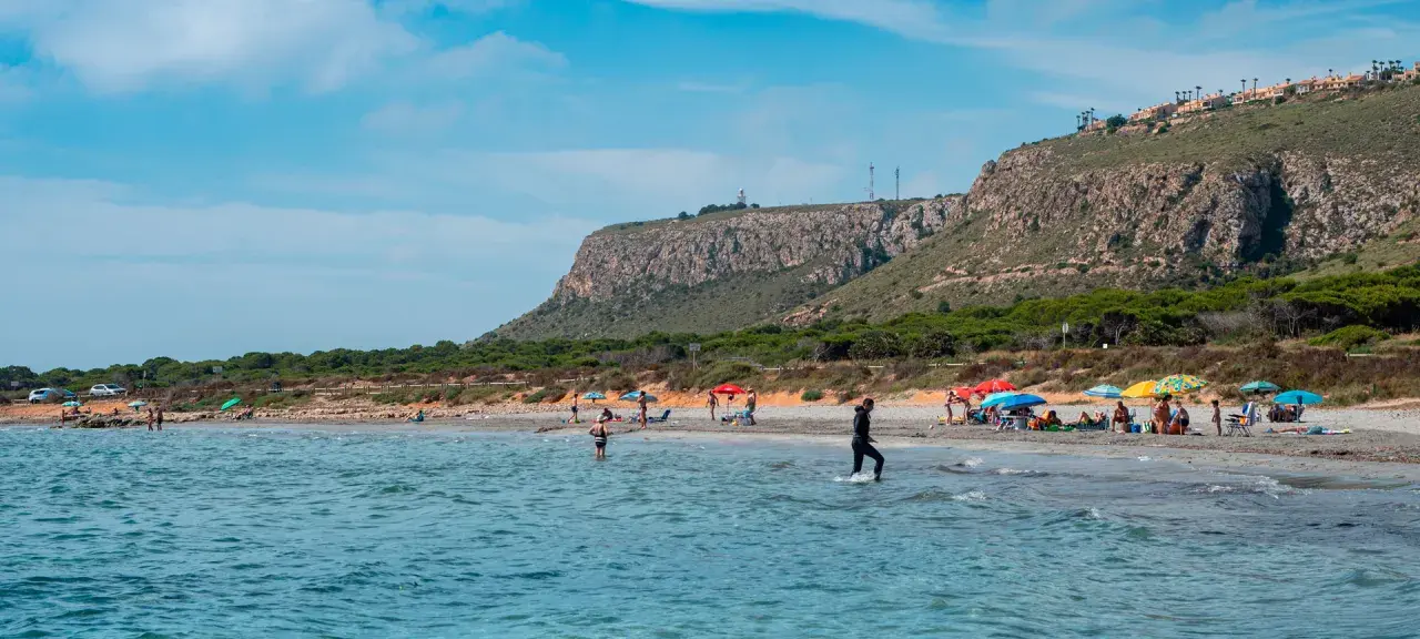 Gente disfruta del sol en la playa de la ermita santa pola, con aguas turquesas y un acantilado verde al fondo.