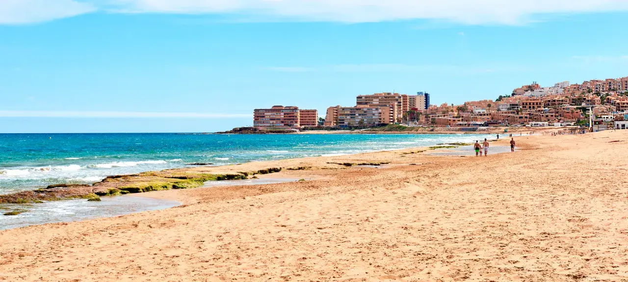 Gente pasea por la **playa de la Mata** con edificios residenciales al fondo y el mar azul.