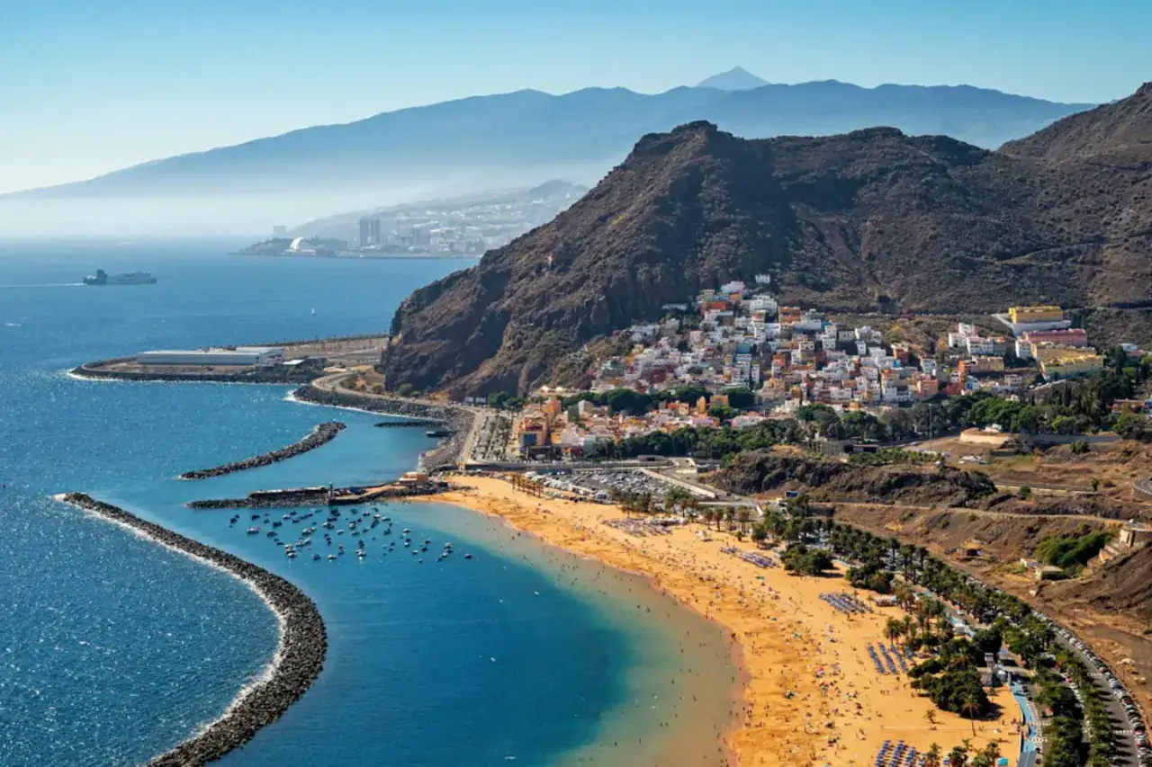 Vista aérea de una playa dorada y un pueblo costero con casas blancas y coloridas en la costa blanca España. Montañas y el mar azul completan la escena.
