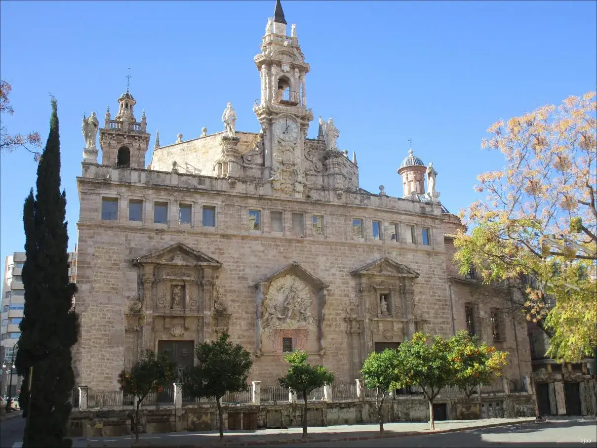 La iglesia de los Santos Juanes, con su imponente fachada de piedra y su torre con reloj, se alza majestuosa bajo un cielo azul.