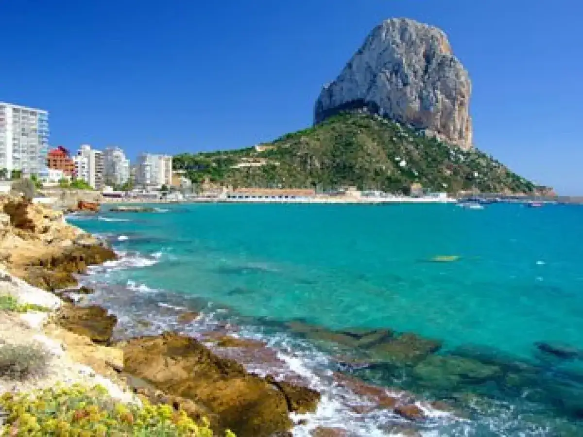 Aguas turquesas y rocas doradas en la mejor playa de Calpe, con el imponente Peñón de Ifach al fondo y edificios blancos a lo lejos.