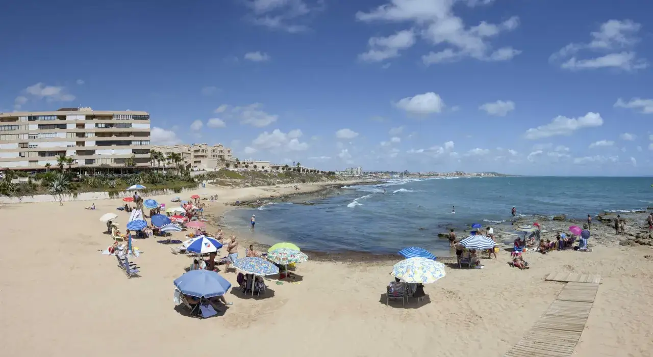 Gente disfrutando del sol y el mar en la playa Cabo Cervera, con sombrillas de colores y edificios al fondo.