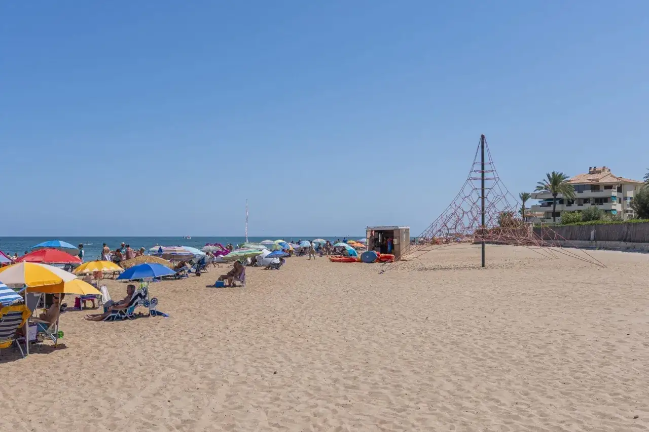 Día soleado en la playa de Les Bovetes, con sombrillas, gente relajándose y una estructura de juegos infantiles.