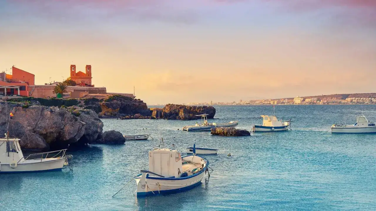 Barcas tranquilas en el puerto de Santa Pola al atardecer, con edificios históricos y la costa al fondo.