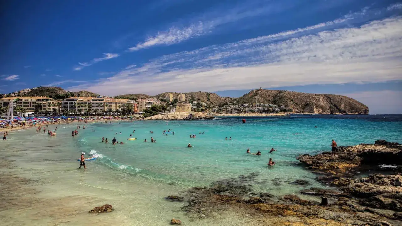 Gente disfrutando del sol y el mar turquesa en la playa de l'Ampolla, Moraira. Edificios y colinas verdes al fondo.