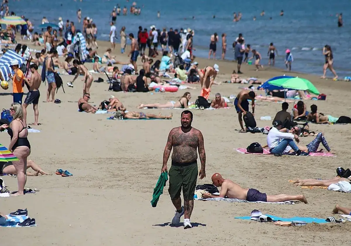 Gente disfrutando del sol en las plages valence. Un hombre con tatuajes camina por la arena mientras otros toman el sol.