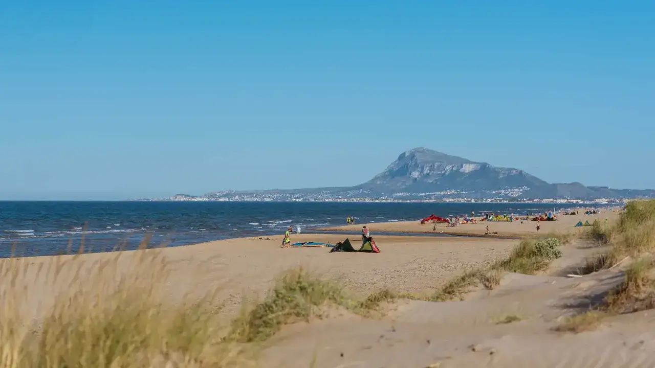 Gente disfrutando del día en la playa de Meliana, con kitesurfistas y tiendas en la arena, bajo un cielo azul y con una montaña al fondo.