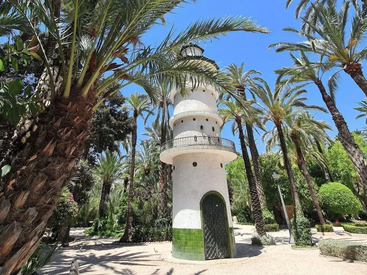 Torre blanca con balcones y puerta verde en el parque el palmeral, rodeada de exuberantes palmeras y vegetación bajo un cielo azul.