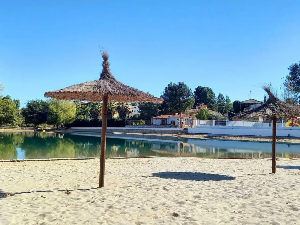 Sombrillas de paja en la playa de Monte Navarrés, con un lago de aguas tranquilas y casas al fondo.