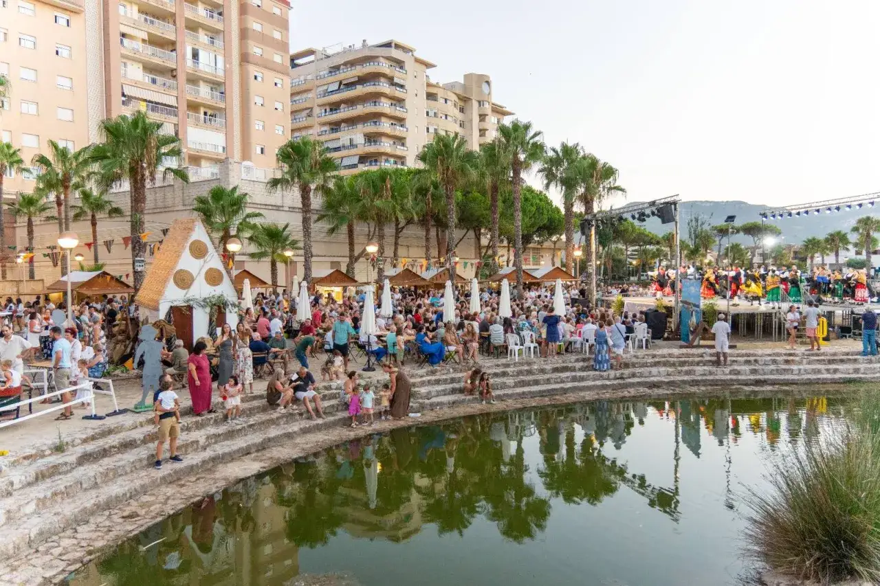 Festival en la playa Tavernes de la Valldigna con puestos, gente sentada en gradas y un escenario con actuaciones.