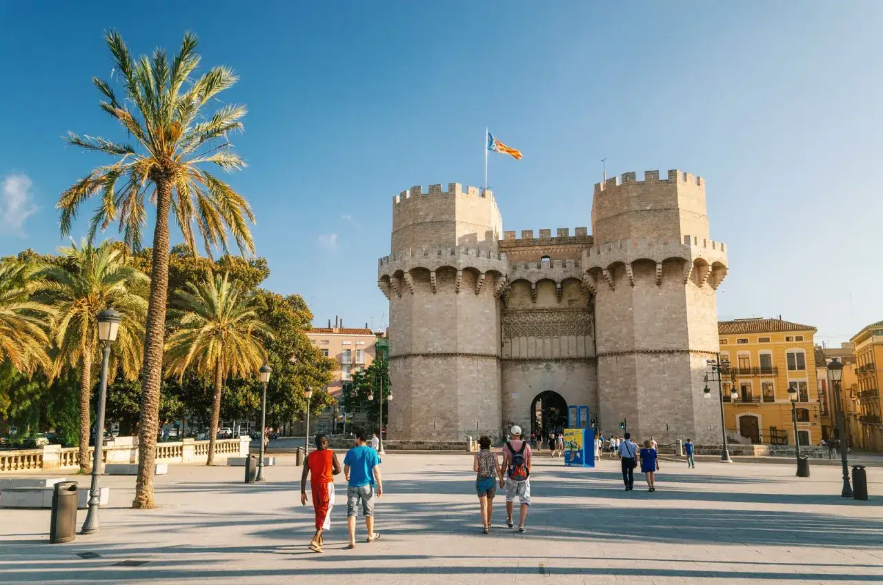 Gente pasea por la plaza frente a las Torres de Serranos, un icono del barrio del Carmen Valencia, bajo un cielo azul y palmeras.