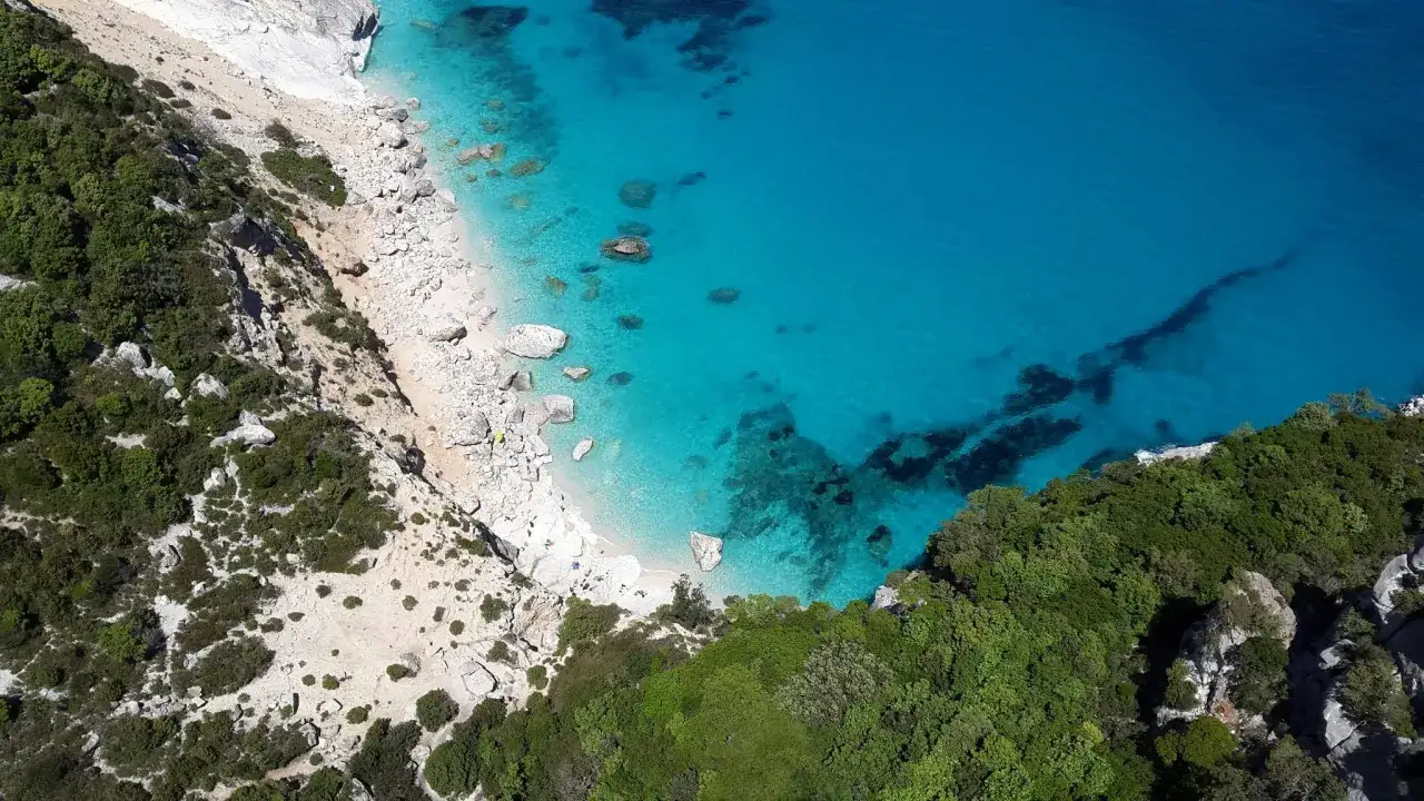 Vista aérea de la playa de Altea, con aguas turquesas cristalinas, arena blanca y rocas, rodeada de vegetación verde y acantilados.