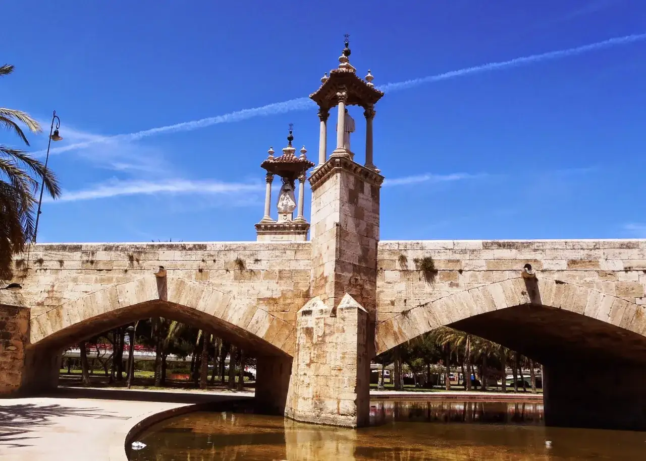 Puente de piedra con estatuas en el Jard&iacute;n del Turia, bajo un cielo azul con estelas de avi&oacute;n.