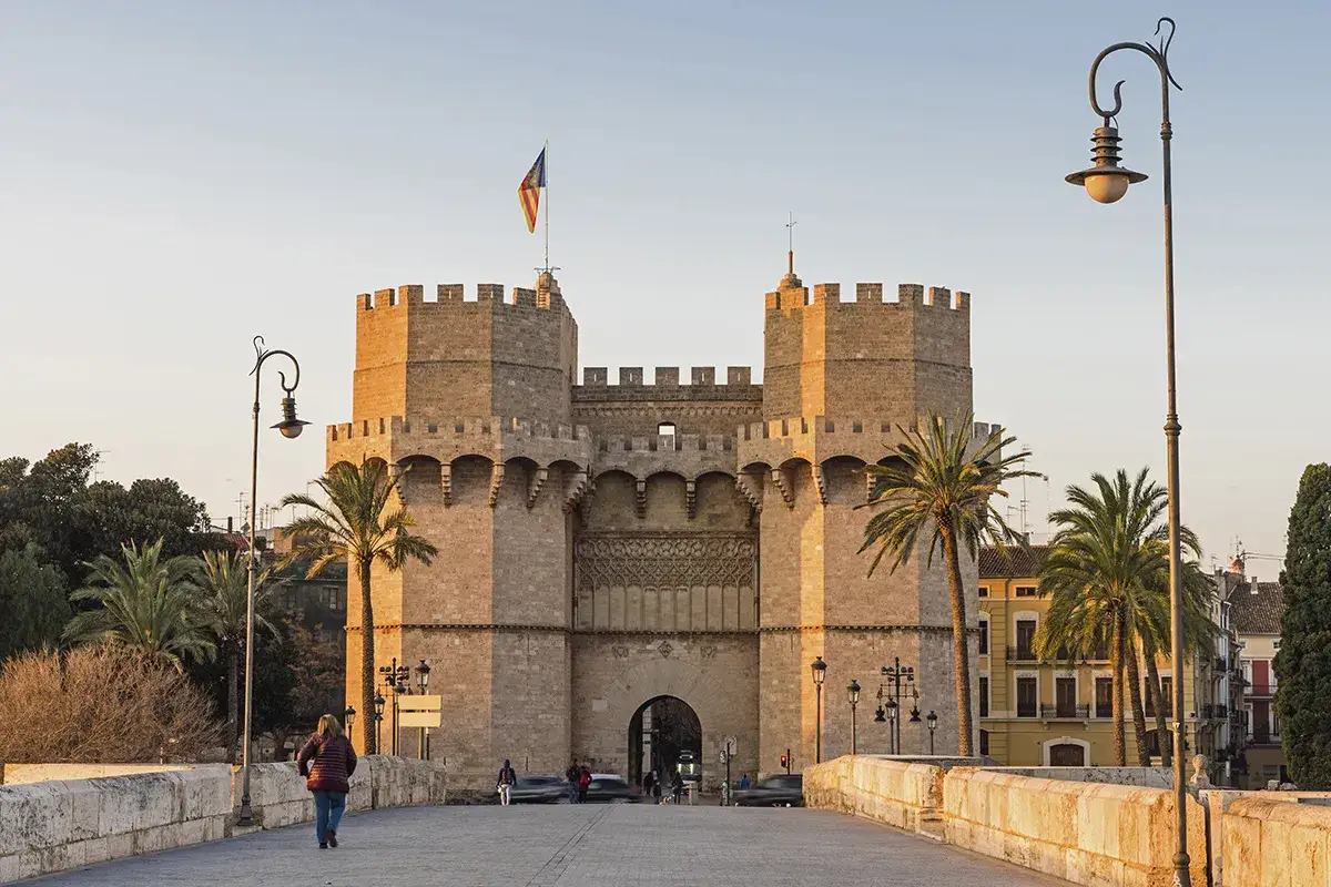 Las Torres de Serranos, imponentes puertas de la antigua muralla de Valencia, se alzan majestuosas bajo un cielo despejado. Palmeras y farolas flanquean el camino.
