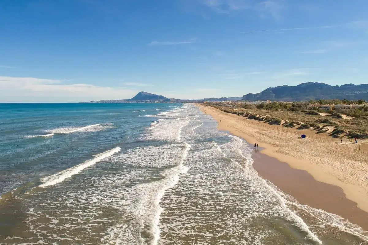 Extensa playa de arena dorada con dunas y el mar Mediterráneo. Al fondo, montañas y el puerto de Oliva, un día soleado.