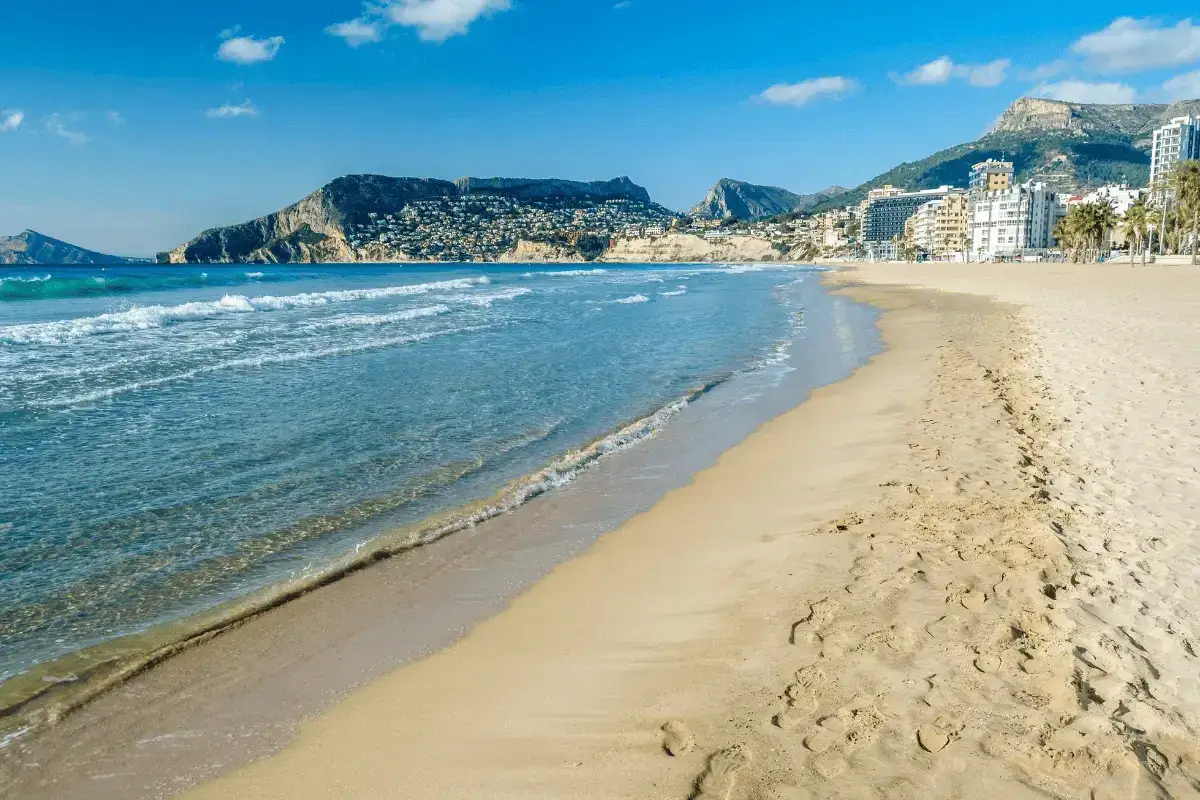 La playa de Calpe, con su extensa arena dorada, invita a pasear junto al mar Mediterráneo. Las olas acarician la costa, mientras el sol ilumina la ciudad y las montañas al fondo.
