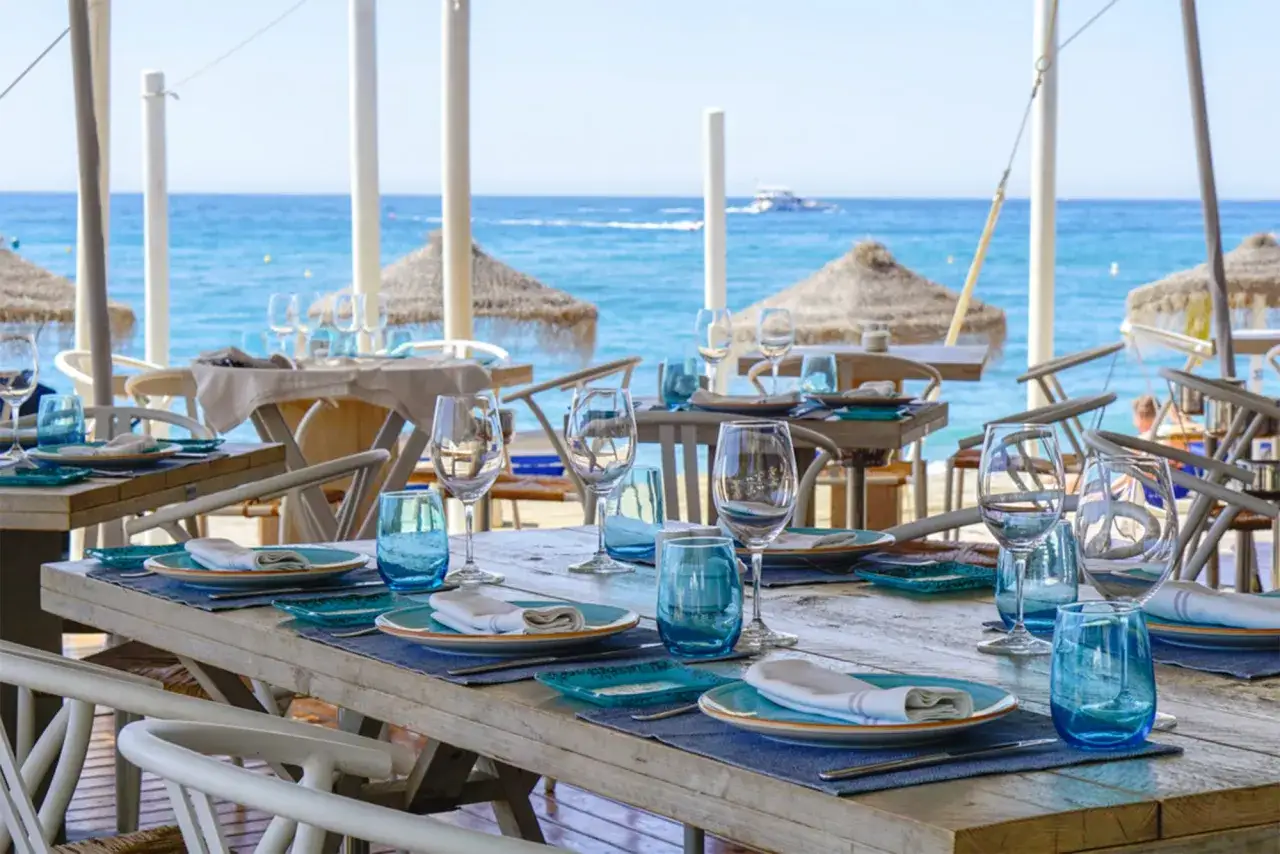 Mesas puestas con vajilla azul y copas de cristal, listas para cenar en la playa de Alicante, con el mar Mediterráneo de fondo.