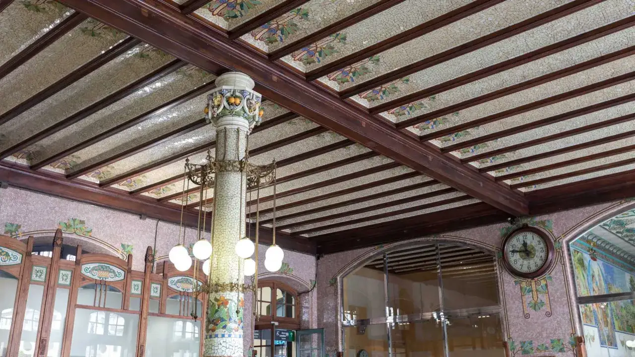 Techo decorado con mosaicos y vigas de madera en la Estación del Norte de Valencia. Una columna modernista soporta lámparas colgantes.