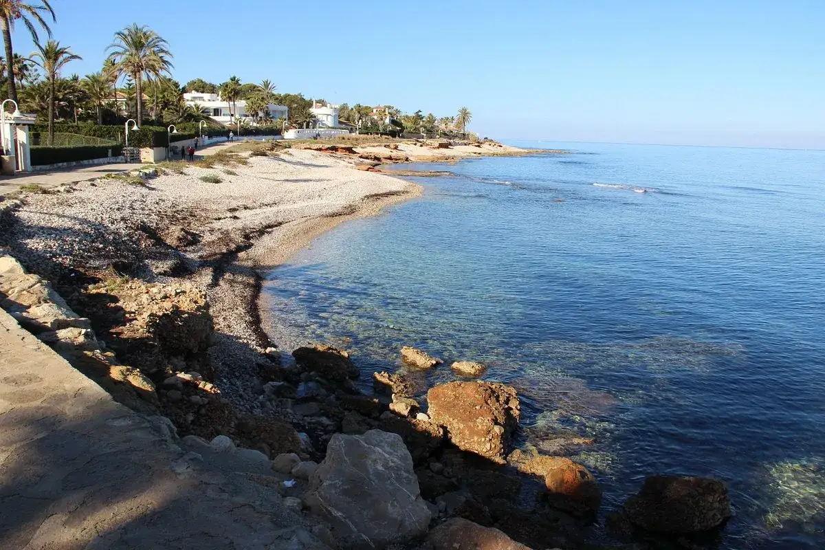 Costa rocosa con playa de guijarros y aguas cristalinas. Palmeras y casas blancas se ven al fondo, cerca de la punta negra.