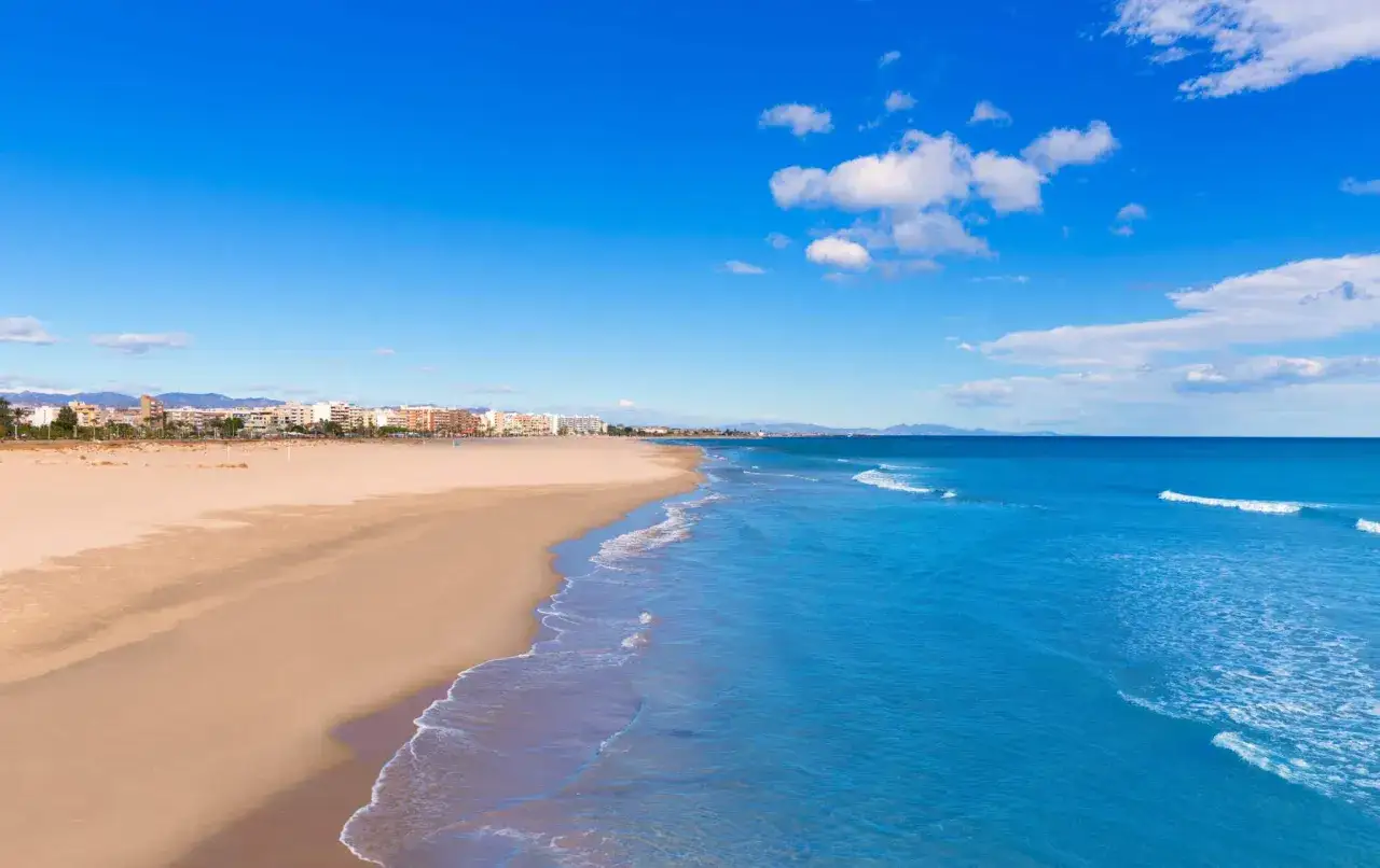 Amplia playa de arena dorada en Sagunto, con el mar azul turquesa y edificios al fondo bajo un cielo despejado.