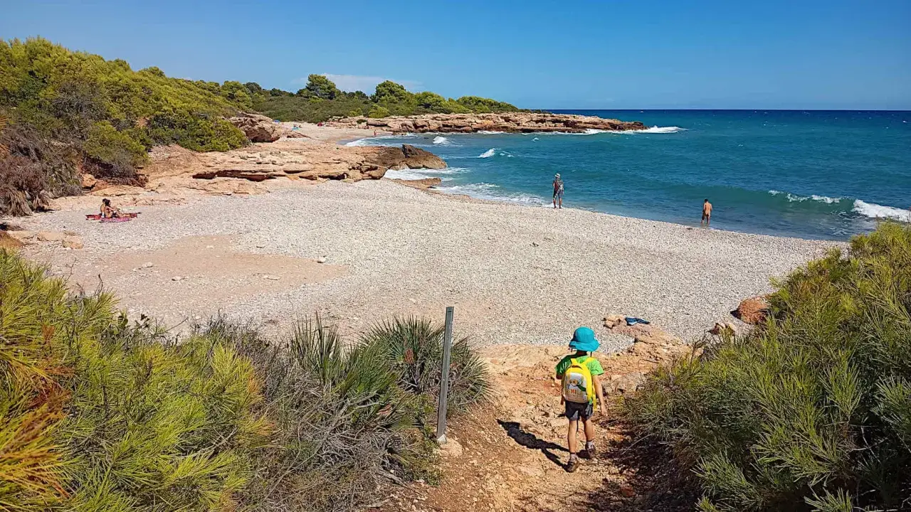 Niño con mochila azul desciende por sendero hacia la cala mundina, de guijarros blancos y aguas turquesas.