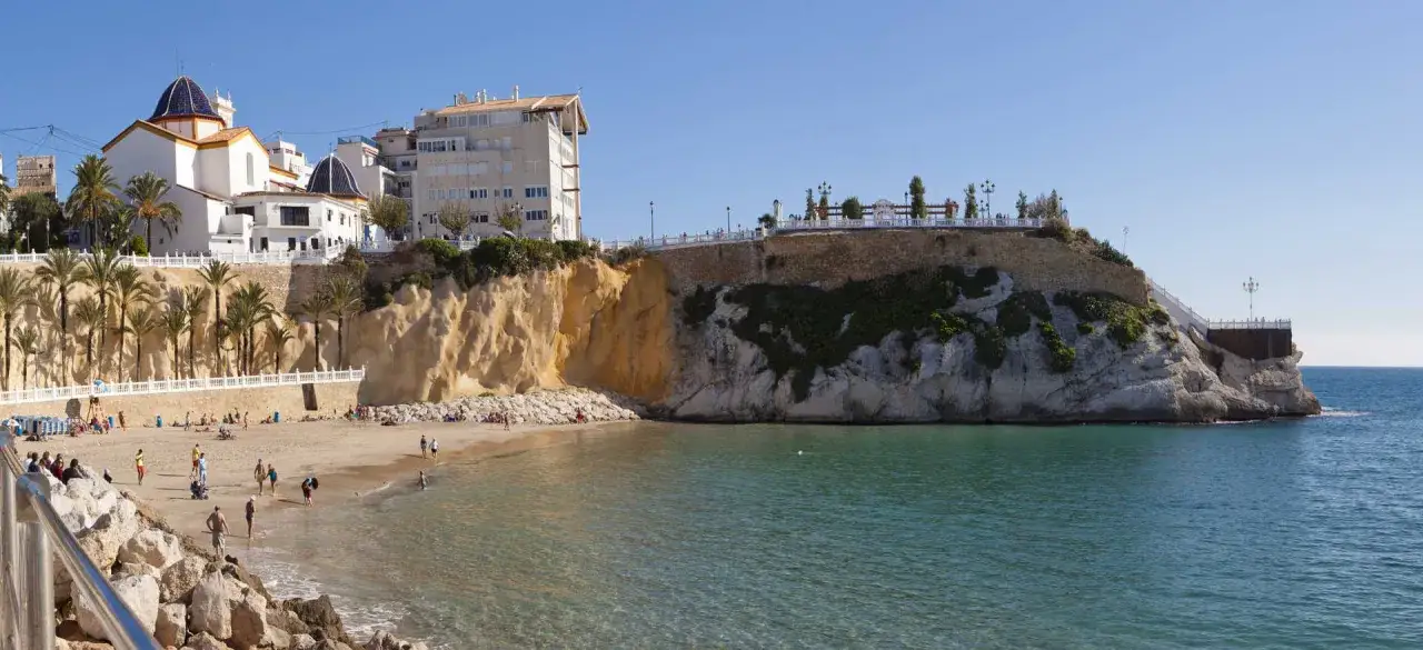Gente disfrutando de la mejor playa de Benidorm, con aguas cristalinas y edificios blancos coronando el acantilado.