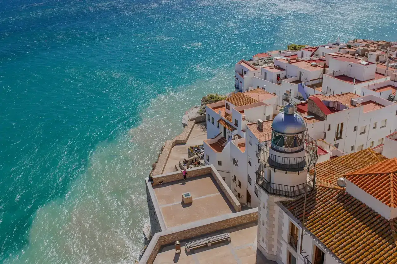 Vista aérea de Peñíscola, con casas blancas y tejados naranjas junto al mar turquesa. El faro se alza imponente, vigilando la playa.