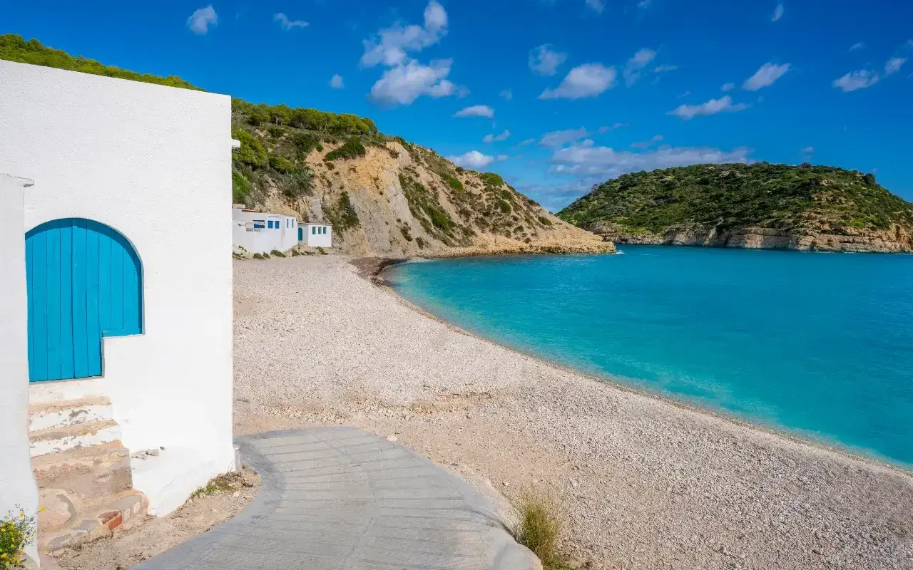 Casa blanca con puerta azul en la playa de la Almadraba, aguas turquesas y colinas verdes.