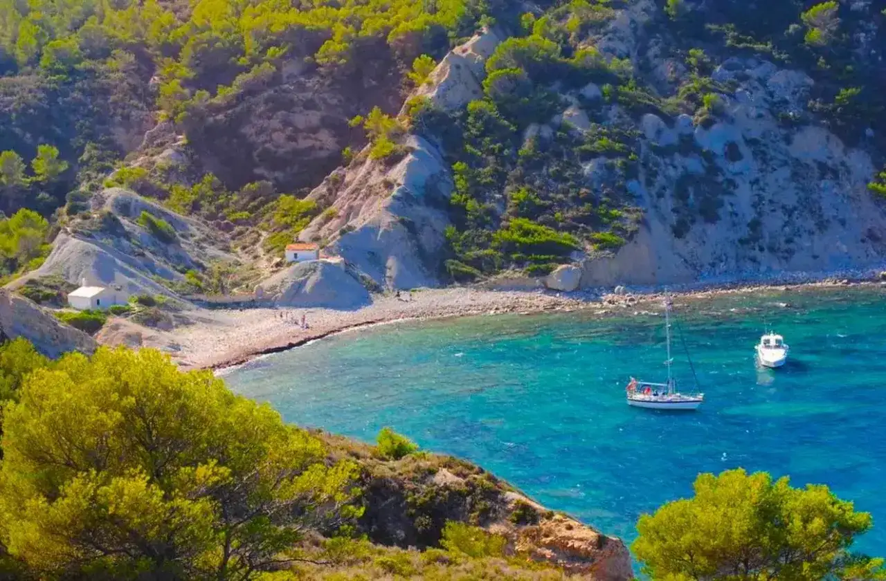 La cala Sardinera, un rincón idílico con aguas turquesas, una playa de guijarros y dos barcos anclados. Dos casitas blancas se asoman entre la vegetación.