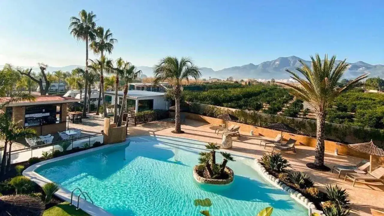 Piscina cristalina y palmeras en un resort para xeraco campers. Caravanas y montañas al fondo.