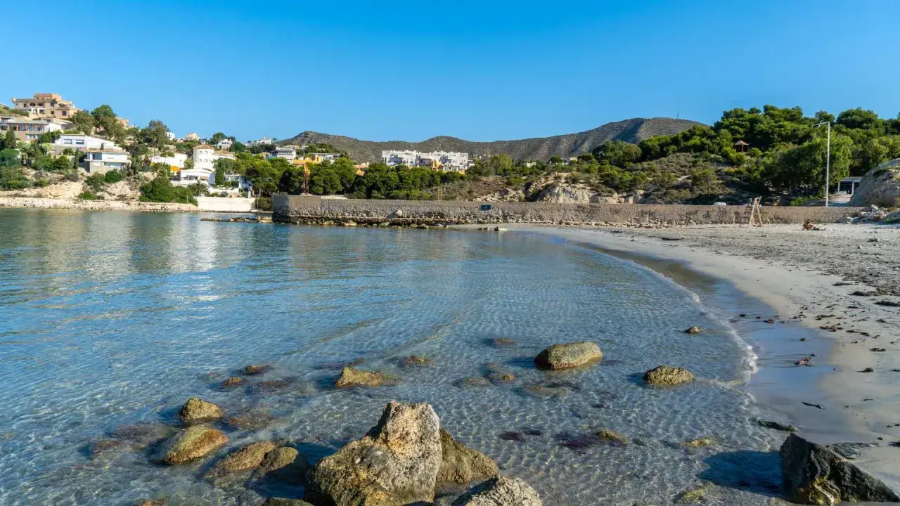 Aguas cristalinas en la cala Morro Blanc, con rocas sumergidas y una playa de arena clara. Al fondo, casas blancas y colinas verdes bajo un cielo azul.