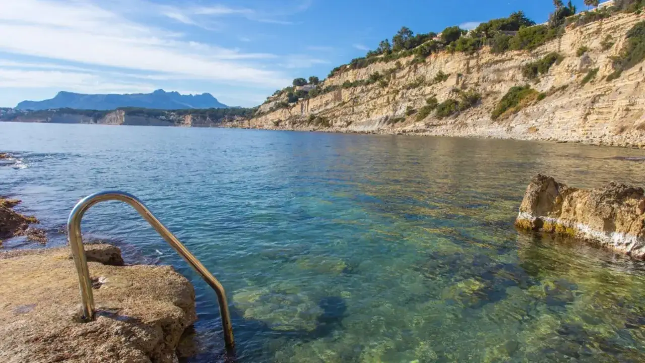 Escalera metálica para acceder a las aguas cristalinas de Punta Estrella Moraira, con acantilados y el mar azul de fondo.