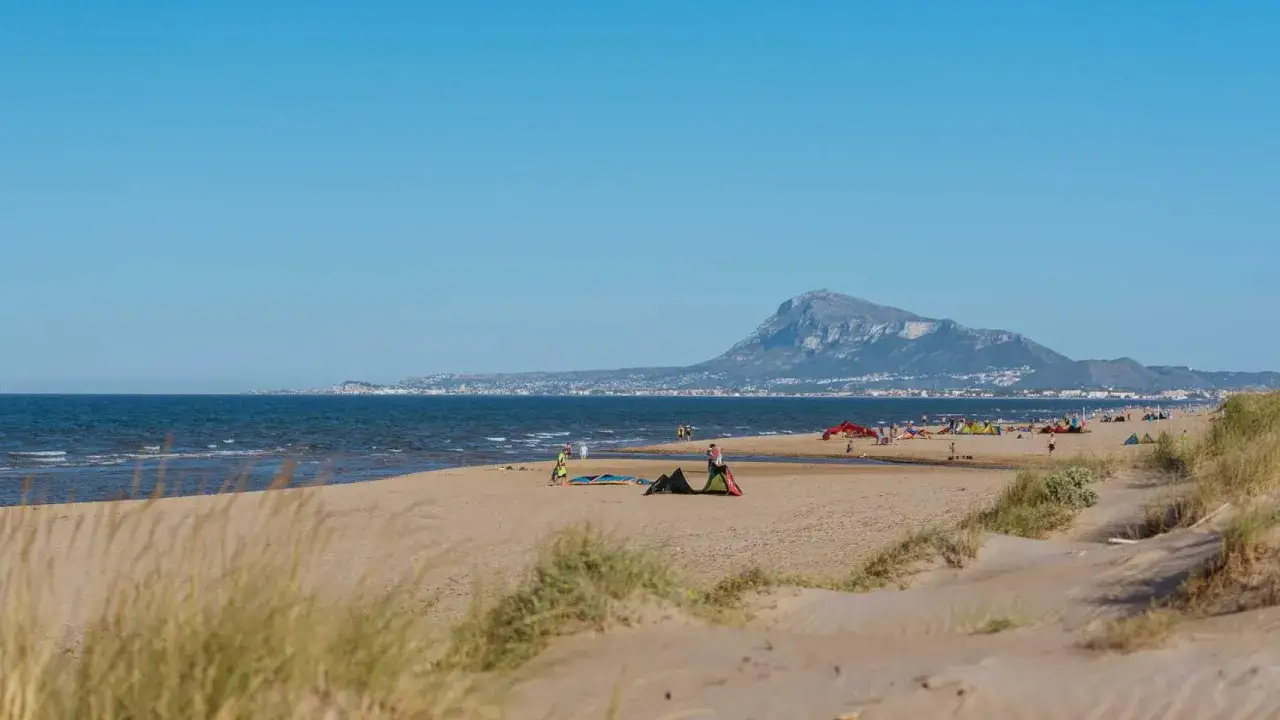 Gente disfrutando de un día soleado en la playa de Rabdells, con el mar azul y una montaña al fondo.