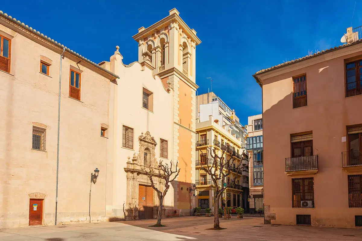 Plaza soleada en la ciutat vella de València, con edificios históricos y un campanario.