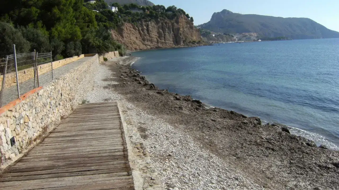 Camino de madera hacia la playa de la solsida, con muro de piedra y acantilados verdes al fondo.