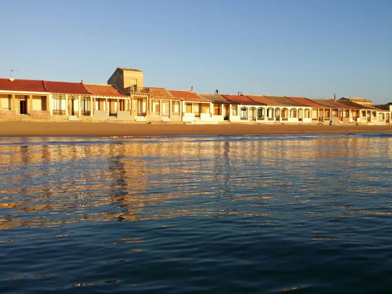 Casitas de colores en la playa Babilonia, reflejándose en el mar azul al atardecer.