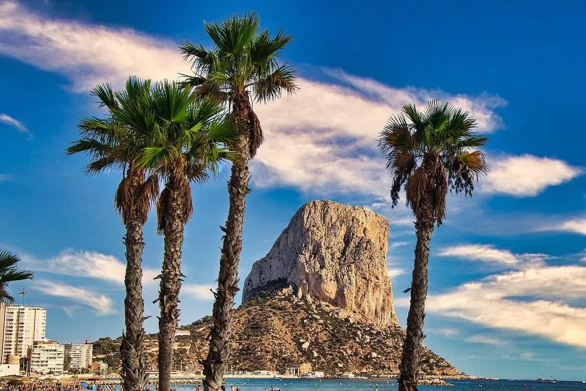 Palmeras en primer plano con el imponente peñón de Calpe al fondo, bajo un cielo azul con nubes blancas.