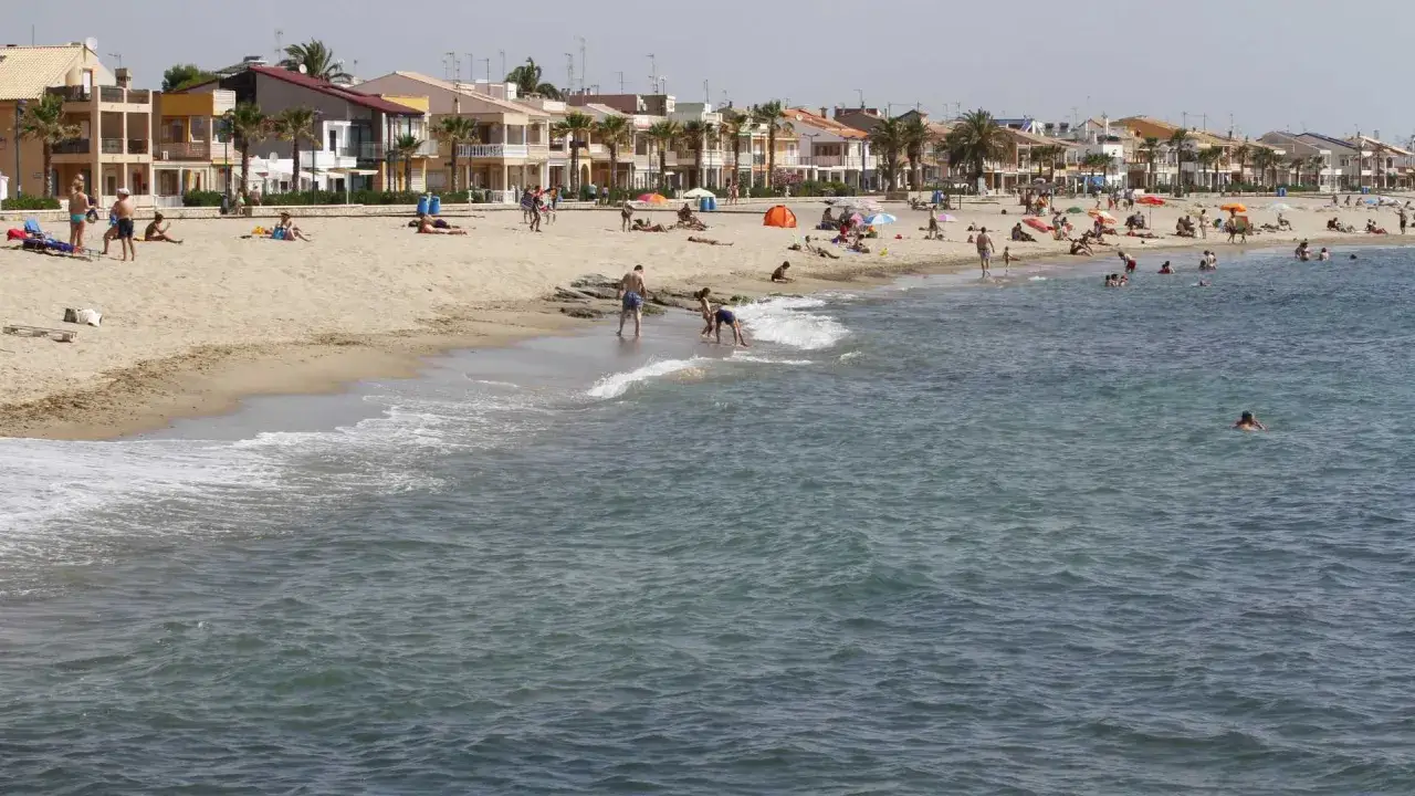 Gente disfrutando del sol y el mar en la playa Puzol, con edificios residenciales al fondo.