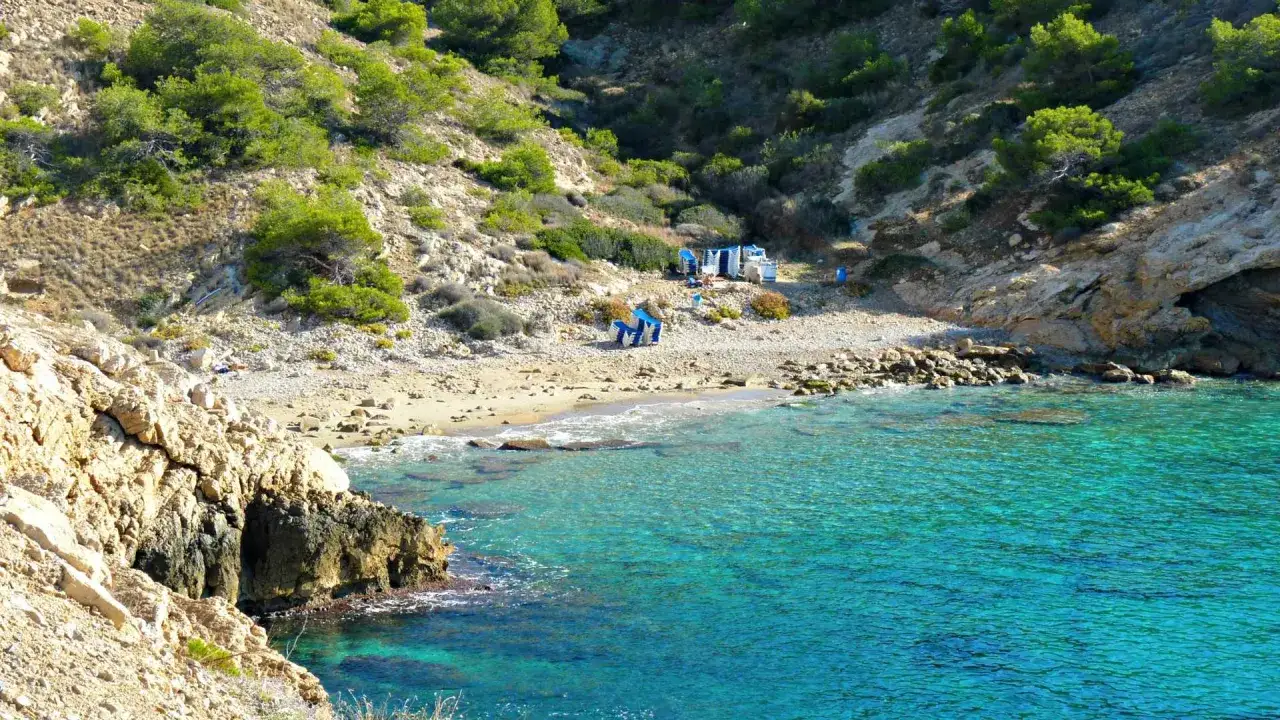 Agua turquesa y rocas doradas en la cala Almadrava, Benidorm. Sombrillas azules y gente disfrutan de este rincón secreto.