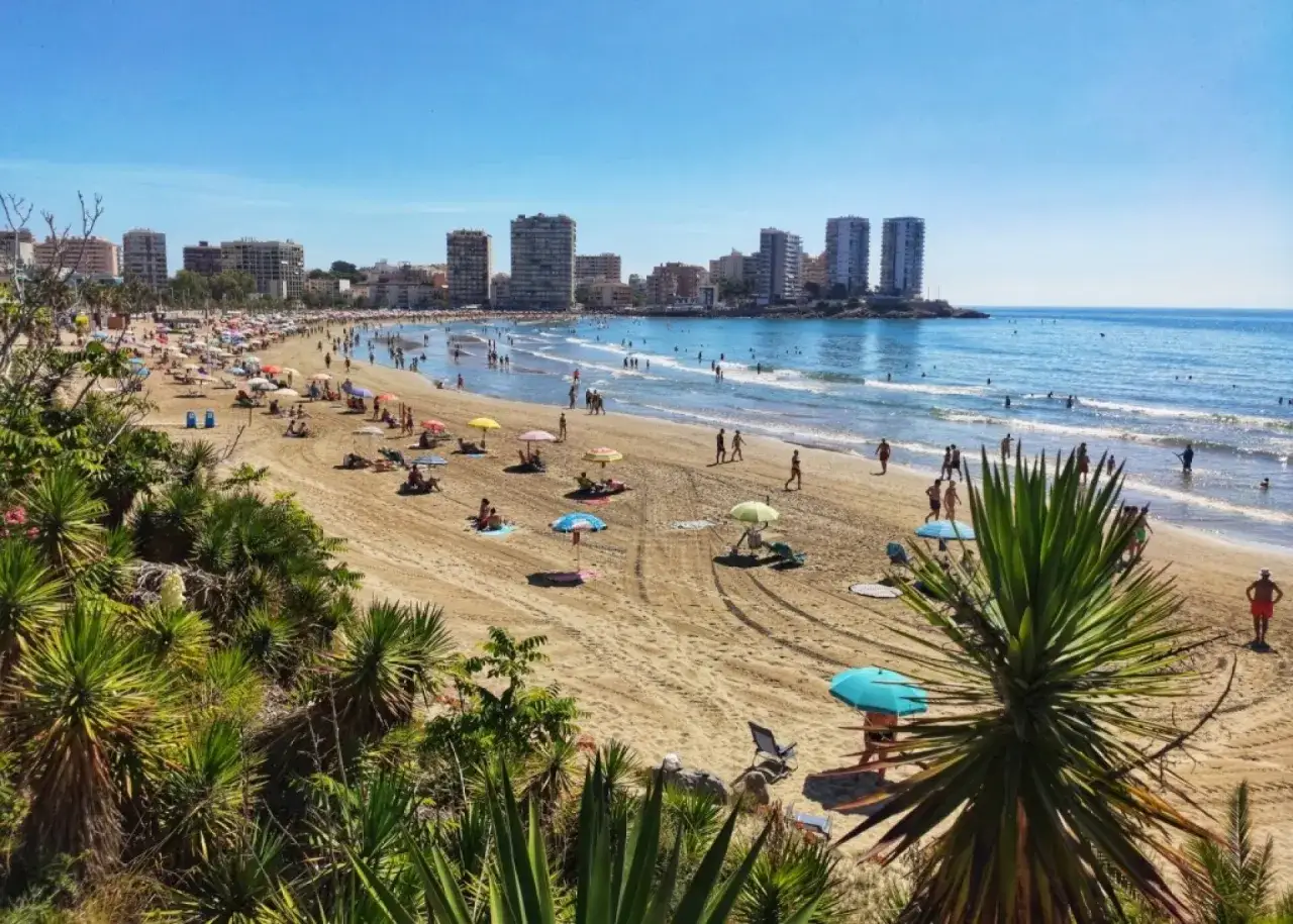 Soleado día en la playa del Moro, con gente disfrutando del mar y la arena bajo sombrillas de colores. Edificios modernos se alzan al fondo.