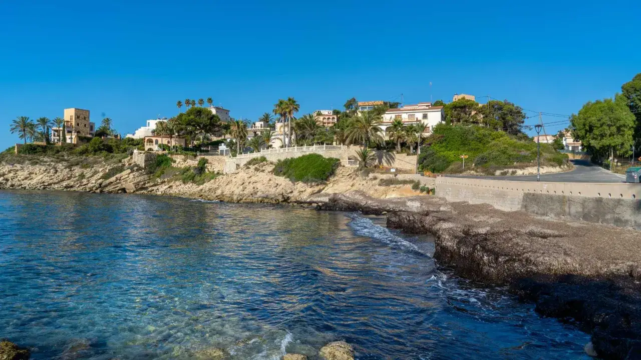 Aguas cristalinas bañan la costa rocosa de cala Piteres, con casas blancas y palmeras coronando el acantilado.
