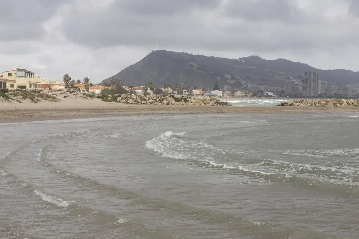 Olas suaves rompen en la playa el Marenyet, con casas y edificios a lo lejos bajo un cielo nublado.