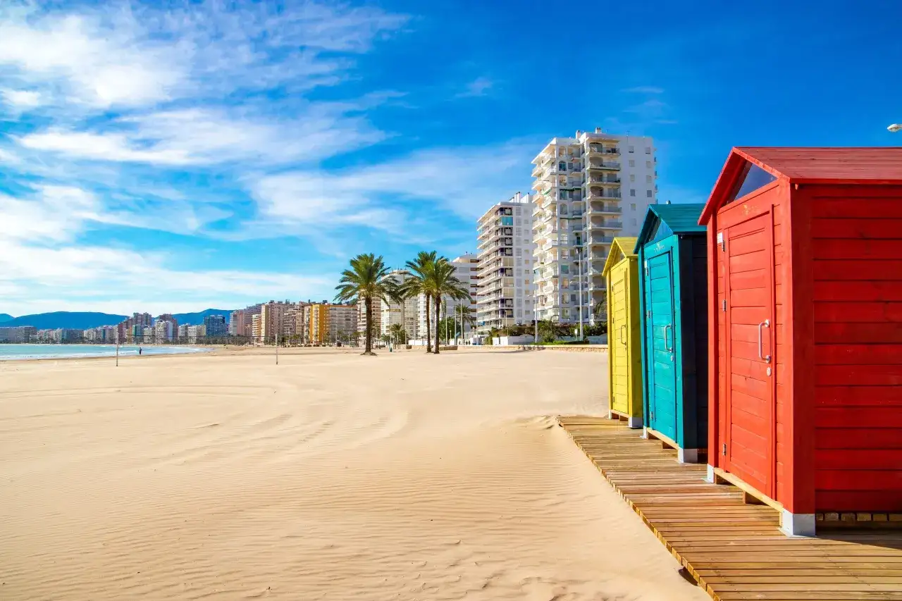 Coloridas casetas de playa en Cullera playa, con edificios altos y palmeras al fondo bajo un cielo azul con nubes.