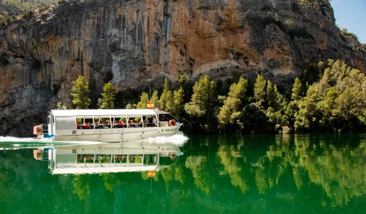 Barco turístico navega por las aguas verdes de los cañones del Júcar, reflejando la imponente pared rocosa y la vegetación.
