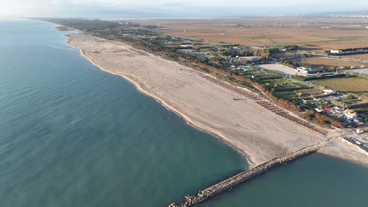 Vista aérea de la playa de Pinedo, con el mar turquesa, arena dorada y un espigón de rocas. Al fondo, campos y edificaciones.