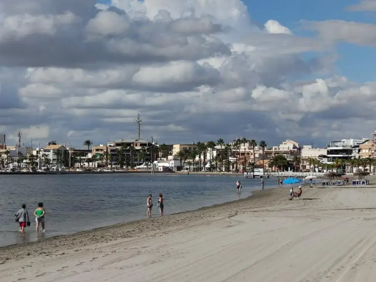 Gente paseando en la playa de las Higuericas, con edificios y palmeras al fondo bajo un cielo nublado.